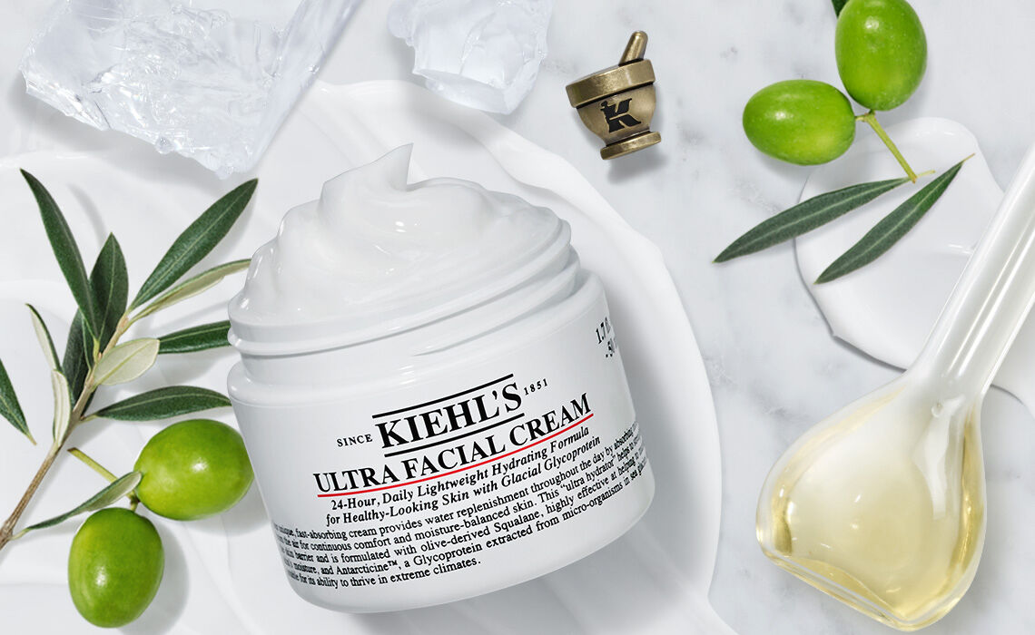 A jar of Kiehl's Ultra Facial Cream surrounded by olive branches, a small bowl of cream, and a decorative spoon on a marble surface.