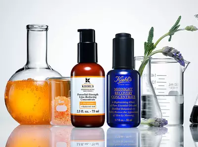 Rozne fľaštičky s kozmetickymi produktmi Kiehl's, obklopene meracimi nadobami a oranžovou kvapalinou.