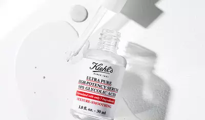 A bottle of Kiehl's Ultra Pure High-Potency Serum containing 38% glycolic acid, accompanied by a droplet of the serum on a white surface. The label emphasizes texture-smoothing benefits.