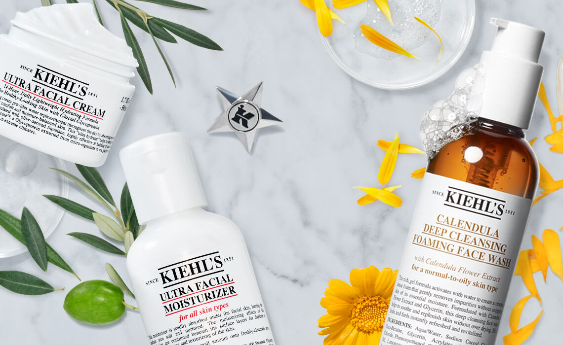 A marble surface featuring Kiehl's skincare products, including Ultra Facial Cream, Ultra Facial Moisturizer, and Calendula Deep Cleansing Foaming Face Wash, surrounded by yellow flowers and green leaves.