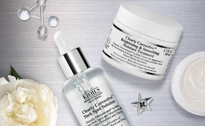 A flat lay of Kiehl's skincare products, including a dropper bottle of Clearly Corrective Dark Spot Solution, a jar of Clearly Corrective Brightening and Smoothing Moisture Treatment, and a white flower, all set against a metallic background.