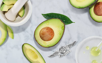 An avocado cut in half, showcasing its seed, surrounded by sliced avocado pieces, green leaves, and a mortar and pestle on a marble surface. The Kiehl's logo is displayed at the bottom right corner.