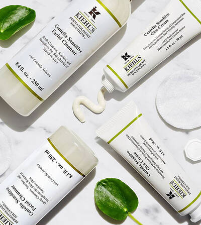 Sada prirodnej kozmetiky Kiehl's vratane čistiaceho gelu a kremu, na bielom pozadi, obklopena zelenymi listami.