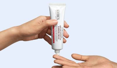 A hand is holding a tube of Kiehl's Ultra Facial Cream, while another hand reaches out to receive the product. The background is light blue.