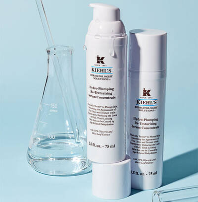 Biele fľaše s pleťovym serum značky Kiehl's na svetlomodrom pozadi s laboratornym sklenenym riadom.