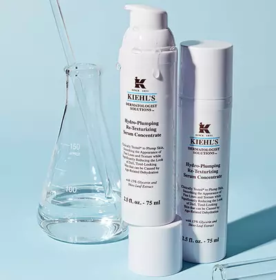 Biele fľaše s pleťovym serum značky Kiehl's na svetlomodrom pozadi s laboratornym sklenenym riadom.