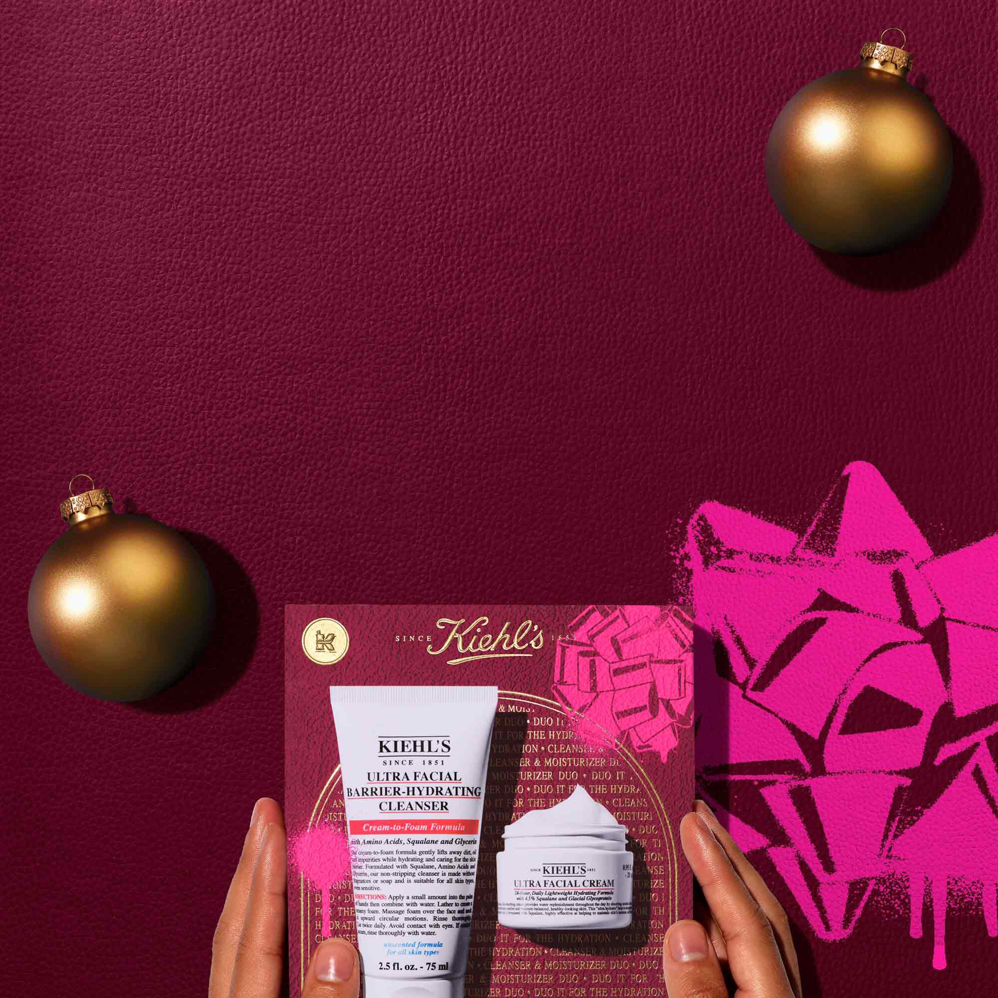 A top-down view of a white tube of Kiehl’s Ultra Facial Barrier-Hydrating Cleanser and a white jar of Kiehl’s Ultra Facial Cream, placed alongside a gift box that features an illustration of the same products. Two golden Christmas ornaments are also visible, one in the top right and one in the bottom left. The gift box in the lower center has a prominent pink graphic of a bow on its right side.