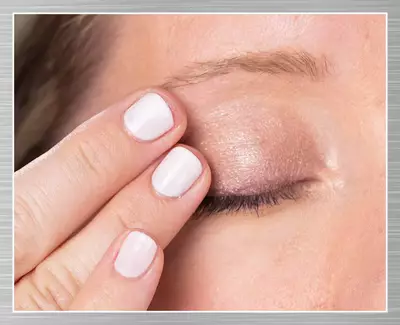 A close-up of a woman’s eye with a finger gently touching her eyelid, showcasing a subtle eyeshadow look. The nails are painted white.