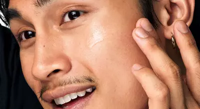 A close-up of a young man applying skincare product to his cheek, smiling, with a slight mustache and wearing an earring.