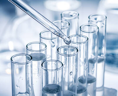 A dropper releasing liquid into a row of clear test tubes filled with water or another transparent liquid, set against a softly blurred laboratory background.