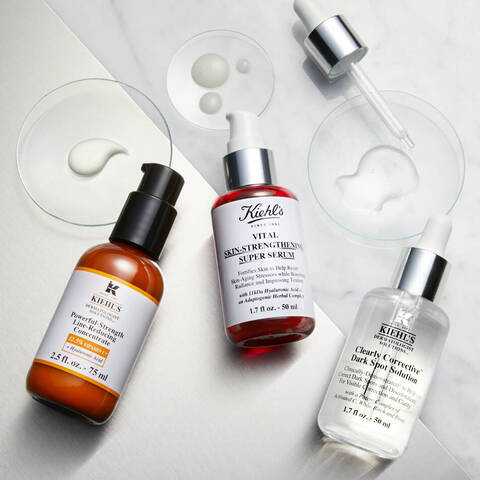 Na obrazku su tri kozmeticke serum značky Kiehl's umiestnene na mramorovej doske, spolu s niektorymi kvapkami produktu v sklenenych miskach.